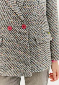 Dvojřadé sako v herringbone vzoru, šedé s multicolor fleky. Obsahuje červené knoflíky a boční kapsu. Měkká textura.