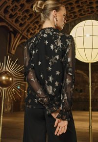 Blusa nera trasparente con motivi di stelle e lune argentate, scollatura e polsini arricciati. Modella in piedi con le mani incrociate dietro la schiena, in un ambiente interno.