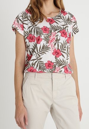 Femme portant un haut blanc à manches courtes avec un imprimé floral rose et oiseaux, associé à un pantalon taille haute beige clair.