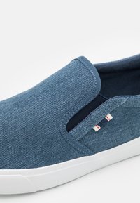 Blauwe denim instapschoen met een witte rubberen zool, gekenmerkt door een textuurstof, gestikte accenten en een klein logo in rood en wit.