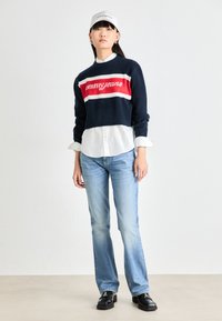 Navy cropped sweater with red and white "omm jeans" logo, layered over a white shirt, paired with light blue flared jeans and black shoes.