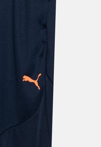 Temno modri športni šorts z teksturirano tkanino. Vključuje oranžno Puma logo na levi strani. Preprost, funkcionalen dizajn.
