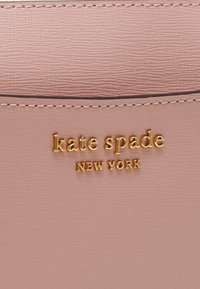 Světlá růžová syntetická kůže s texturovaným povrchem, zdobená zlatým embosovaným logem "kate spade NEW YORK" a kontrastním prošíváním.