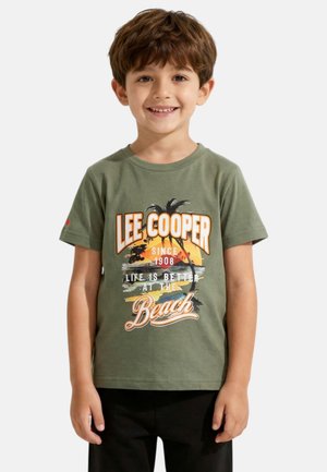 Jeune garçon souriant, portant un T-shirt vert avec un motif de palmier et coucher de soleil sur la plage, et un pantalon noir, debout devant un fond uni.