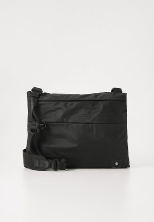 Sort, rektangulær taske lavet af holdbart stof. Har en justerbar rem, hardware på siden og et lille logo tag i nederste hjørne.