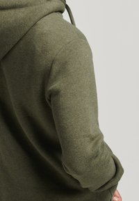 Sudadera verde oliva con una textura suave, cordón ajustable y puños acanalados. Enfoque en el área del hombro y la parte superior del brazo.
