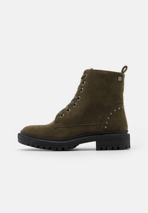 Bottines en daim vert olive avec un bout arrondi, un laçage à l'avant et une semelle en caoutchouc noir. Elles sont ornées de clous argentés sur le talon.