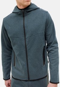 Capuchonjack in donker teal, gemaakt van textuurstof. Voorzien van een ritssluiting aan de voorkant, zijzakken en contrasterende zwarte ritsen en afwerking.