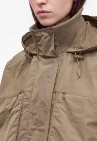 Gros plan d'une personne portant une veste taupe avec un col côtelé haut, une capuche, un bouton pression et des cordons réglables.