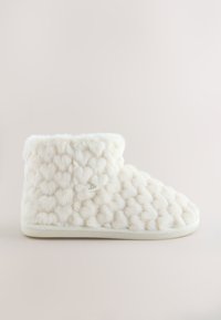Chausson-botte en peluche blanche avec motif en forme de cœur texturé et semelle souple, présenté de profil sur un fond uni.