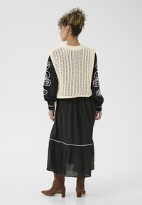 Jersey crema sobre un vestido negro; el vestido presenta un borde blanco y un dobladillo voluminoso. La modelo lleva botas de ante marrón hasta el tobillo.