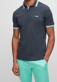 Man som bär en mörk marinblå polo med vit och turkos kant och "BOSS"-logotyp, tillsammans med ljus turkosa byxor, med en hand i fickan.