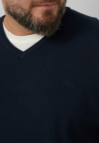 Granatowy sweter w serek wykonany z dzianiny, z delikatnym logo wytłoczonym na klatce piersiowej, noszony na białej koszulce.