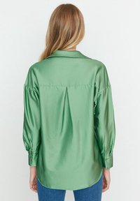 Grön satin skjorta med krage, långa puffärmar och knappslut, med en något rundad nederkant och detaljer med veck bak.