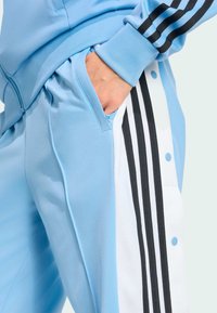 Light blue athletic tracksuit pants featuring a side pocket, with black and white stripes along the leg and snap buttons for adjustment.
