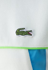 Polo shirt texturé blanc avec le logo crocodile vert de Lacoste. Présente des accents bleus et des bordures vertes le long des coutures pour le contraste.