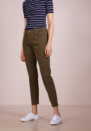 Personne portant un pantalon slim vert olive, un t-shirt rayé bleu marine et blanc, et des baskets blanches, debout devant un fond clair uni.