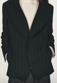 Czarny marynarka w prążki o luźnym kroju, wykonana z fakturowanej tkaniny, bez klap i z bocznymi kieszeniami, noszona na beżowym swetrze.