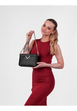 Black leather handbag with a glossy finish, V-shaped accent, and gold chain strap. Held by a person wearing a red dress.