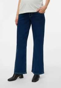 Plaša kāju tumši zilās džinsu bikses ar gludu tekstūru un kontrastējošu šuvju, apvienotas ar melniem blokveida papēžiem līdz potītei.