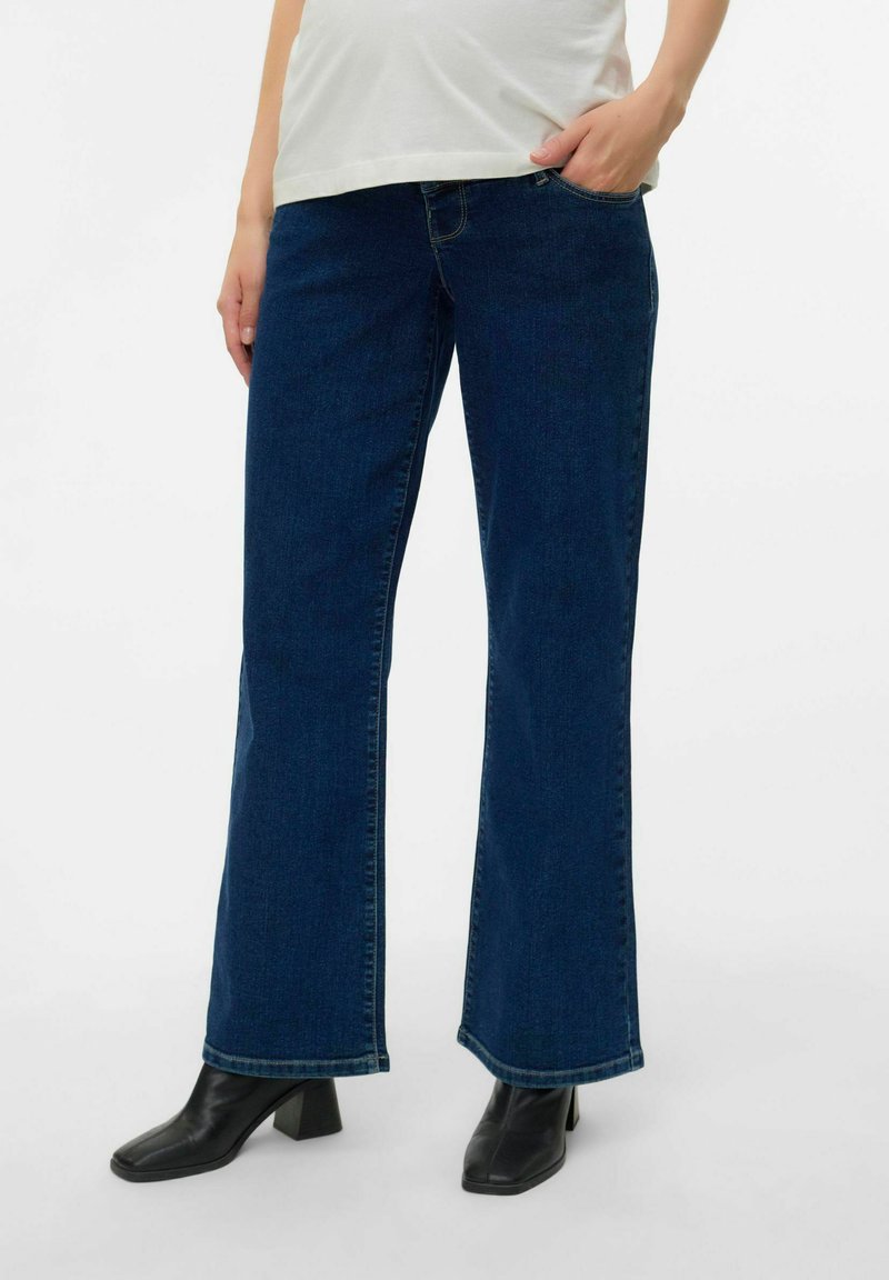 Plaša kāju tumši zilās džinsu bikses ar gludu tekstūru un kontrastējošu šuvju, apvienotas ar melniem blokveida papēžiem līdz potītei.