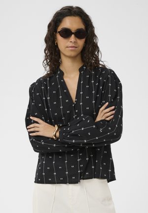 Žena v čiernej blúzke s potlačou a nafúknutými rukávmi, svetlých nohaviciach, slnečných okuliaroch a zlatom náramku, so skríženými rukami na bielom pozadí.