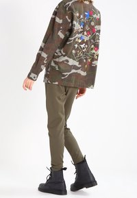 Kamouflage-mönstrad jacka i brunt och grönt med blommig broderi på ryggen, i kombination med olivgröna byxor och svarta snörkängor.