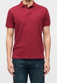 Homme portant un polo bordeaux à manches courtes avec un petit logo vert sur la poitrine gauche et un jean bleu foncé.
