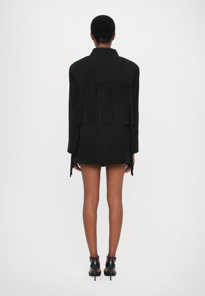 Giacca in suede nera con dettagli in frange sul retro, abbinata a una minigonna. Il modello indossa stivaletti neri alla caviglia.