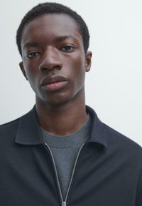 Mørk lynlåstrøje med høj krave, lagdelt over en grå trøje. Glat tekstur og minimalistisk design mod en lys baggrund.