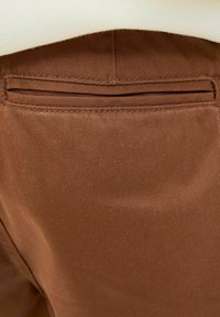 Tissu brun avec une texture lisse et une ouverture de poche cousue avec deux plis discrets. Détails cohérents avec le design de pantalons tailleur.