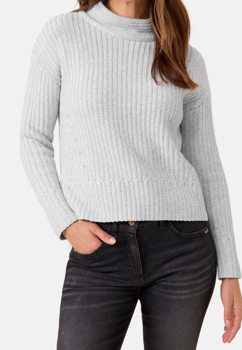 Femme portant un pull en maille côtelée gris clair et un jean noir, la main droite dans la poche et les cheveux lâchés sur l'épaule.