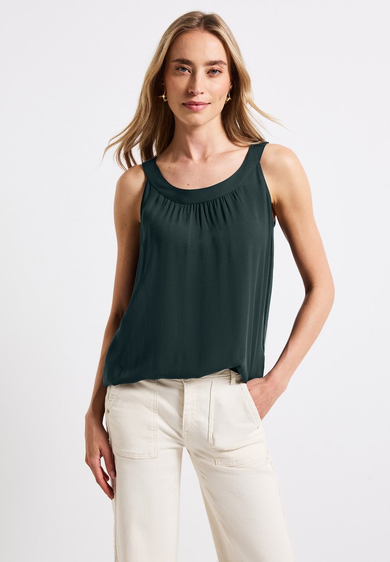 Top verde senza maniche con scollatura rotonda e dettagli arricciati. Tessuto morbido che scivola liberamente. Abbinato a pantaloni larghi color crema.