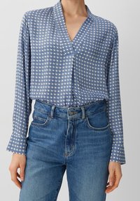 Blå mönstrad blus med lång ärm, med en V-ringning och en geometrisk design. Matchad med högmidjade jeans.