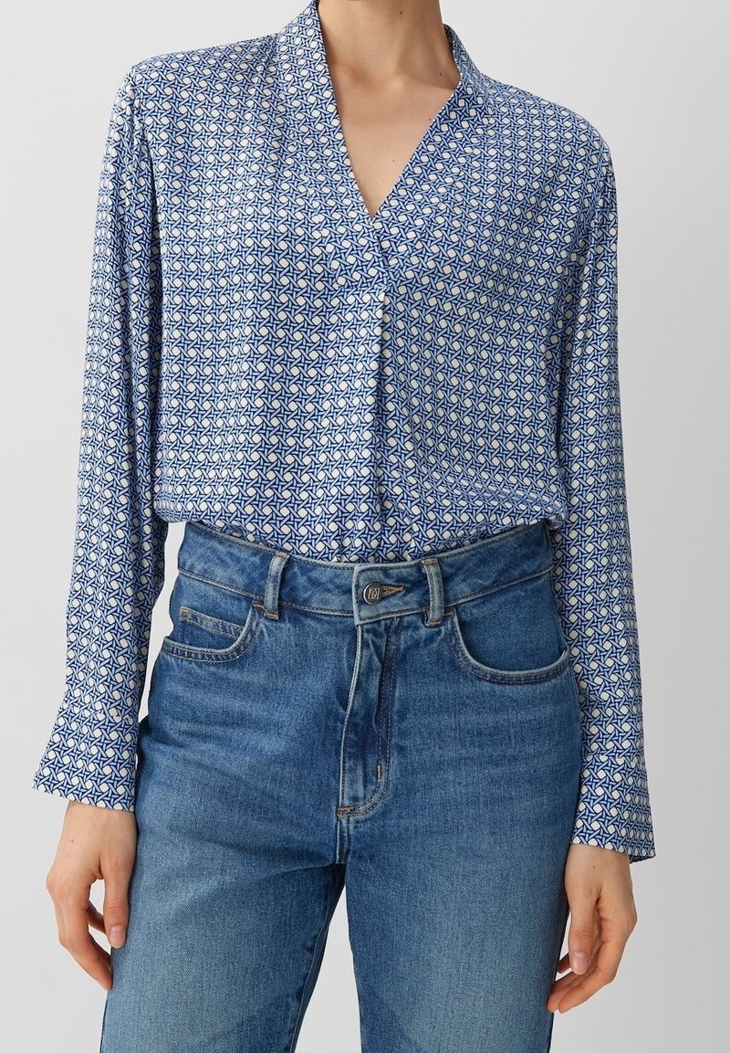 Blå mönstrad blus med lång ärm, med en V-ringning och en geometrisk design. Matchad med högmidjade jeans.