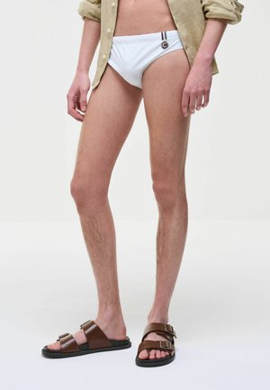 Osoba w białych slipach kąpielowych z logo, beżowej, podwiniętej koszuli oraz brązowych sandałach z klamrą, stojąca na jednolitym białym tle.