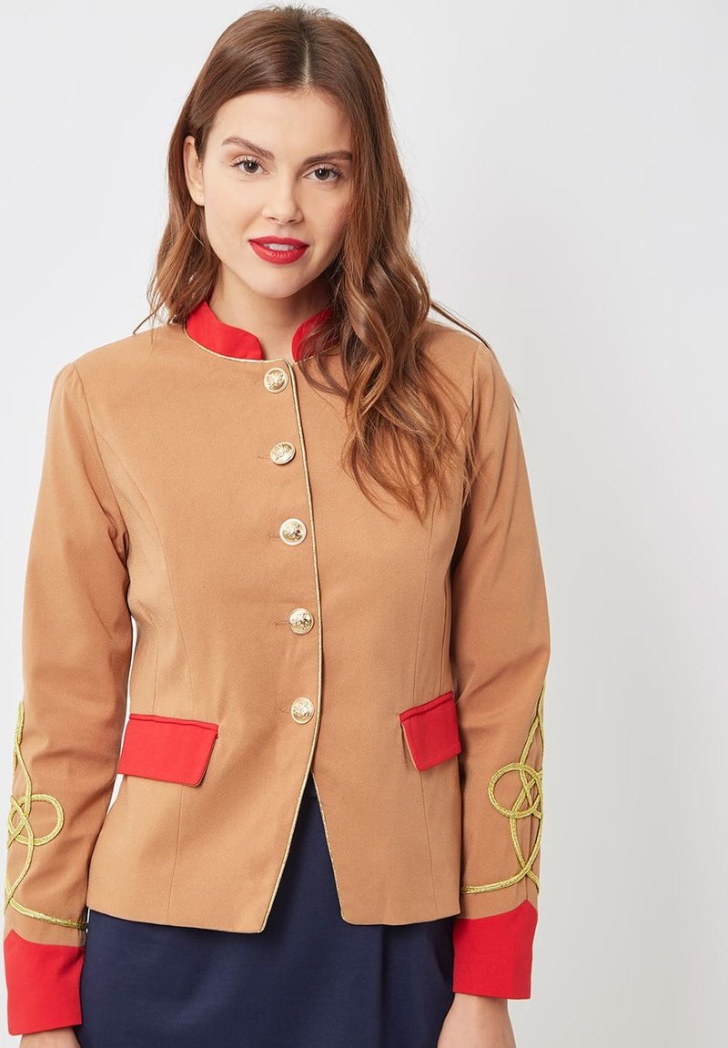 Mujer viste una chaqueta marrón claro con cuello rojo, puños, solapas de bolsillo, botones dorados y diseños dorados bordados en las mangas.