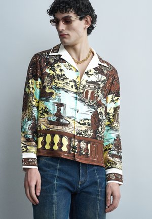 Joven con cabello rizado que lleva una camisa de manga larga con estampados de fuentes, estatuas y escenas de jardines, combinada con jeans azules y una cadena dorada.