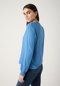 Hellblaues Langarmshirt mit lockerer Passform, ausgestattet mit gerafften Bündchen und weichem Material. Getragen mit dunklen Jeans.