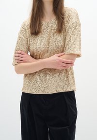 Femme aux longs cheveux bruns portant un haut doré à sequins à manches courtes et un pantalon noir, les bras croisés sur la poitrine devant un fond uni.