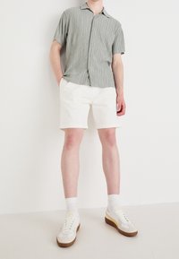 Camicia a maniche corte a righe verdi con bottoni, abbinata a pantaloni corti bianchi e sneakers bianche con accenti gialli e suole marroni.
