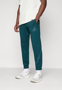 Teal joggingbukser lavet af blødt stof, med en tapered fit, lynlås lomme og en subtil Adidas-logo i en lysere nuance. Sammen med lyse sneakers.