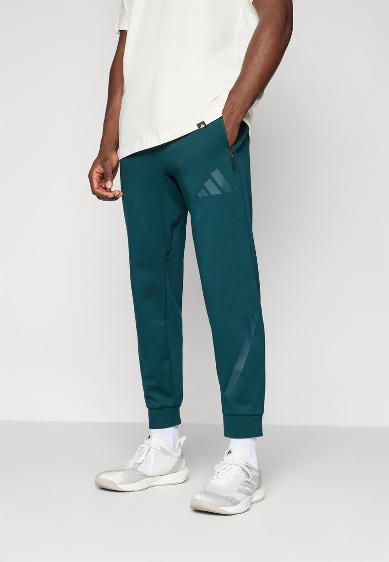 Teal joggingbukser lavet af blødt stof, med en tapered fit, lynlås lomme og en subtil Adidas-logo i en lysere nuance. Sammen med lyse sneakers.