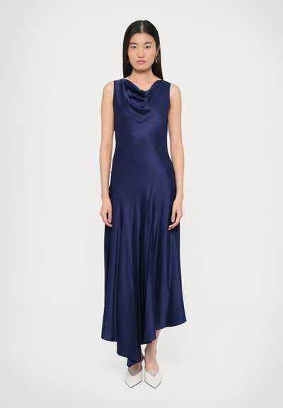 Robe en satin bleu marine avec un col bénitier, un design sans manches, un ourlet fluide et une longueur asymétrique, associée à des chaussures blanches à bout pointu.