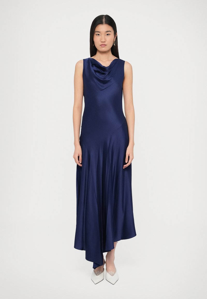 Tengerészkék szatén ruha lejtsző nyakkivágással, ujjatlan kivitelben, légies szegéllyel és egyenetlen hosszúsággal, fehér hegyes cipőkkel párosítva.