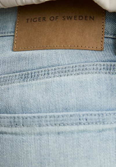 Lyseblå denim med synlig søm og et brunt lærpatch med pregede bokstaver "TIGER OF SWEDEN." Glatt tekstur, uformell design.