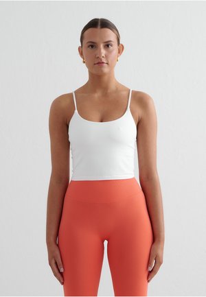 Frau mit dunklen Haaren, die ein weißes Tanktop und korallene Leggings mit hoher Taille trägt, vor einem schlichten hellen Hintergrund steht.