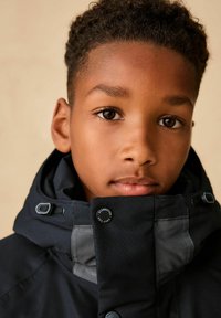 Schwarze Kapuzenjacke mit grauen Akzenten, ausgestattet mit einem hohen Kragen, Knopfverschluss und Kordelzug-Details. Das Material wirkt glatt und strapazierfähig.