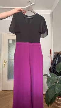 Mano che tiene un appendiabiti con un vestito caratterizzato da maniche corte trasparenti nere e scollatura a V, pantaloni larghi magenta in una stanza con una pianta e un attaccapanni.
