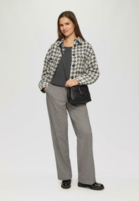 Zwart-wit houndstooth jas, grijze top, grijze broeken en een kleine zwarte handtas. Het model draagt glanzende zwarte schoenen en heeft een ontspannen pasvorm.